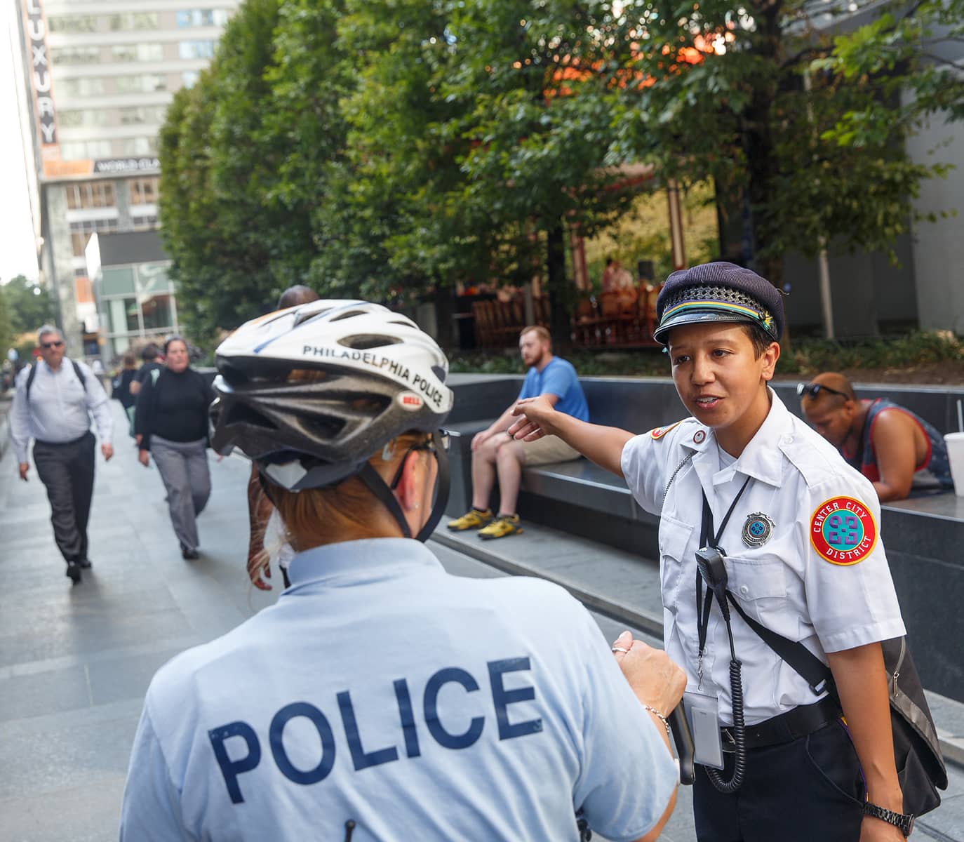 Police officers engage in a conversation on a city sidewalk. One gestures to the side. People walk by and sit on benches amid urban greenery.