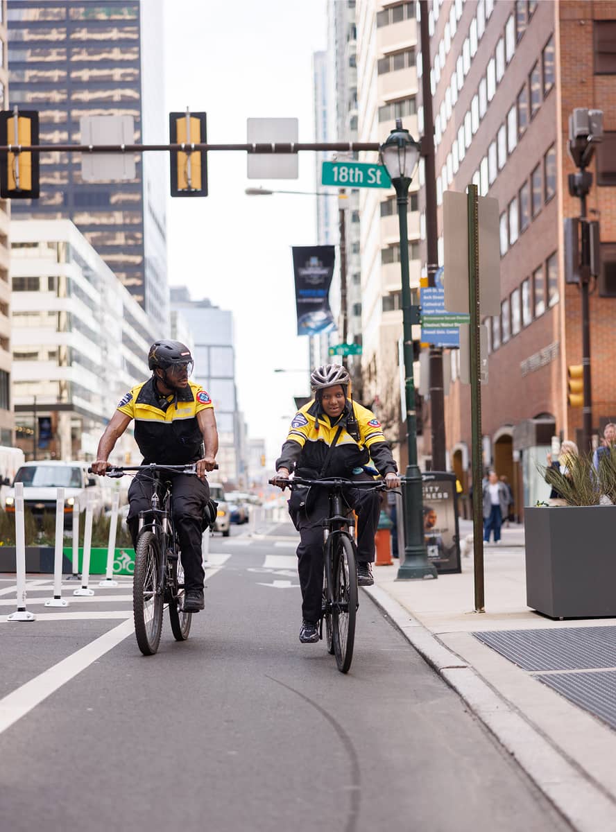 Two uniformed security personnel ride bicycles down a city street, lined with tall buildings. They wear helmets and appear focused and alert.