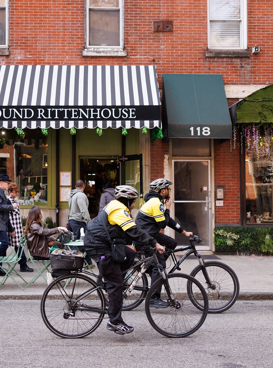 Two cyclists in yellow jackets ride past a brick building with a striped awning. A few pedestrians and a seated person are visible, conveying an urban scene.