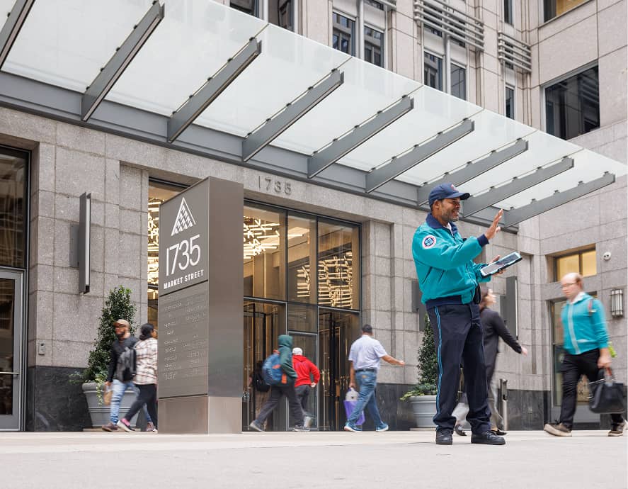 A security officer in uniform directs pedestrians in front of the modern entrance of 1735 Market Street, a tall office building with large glass doors.