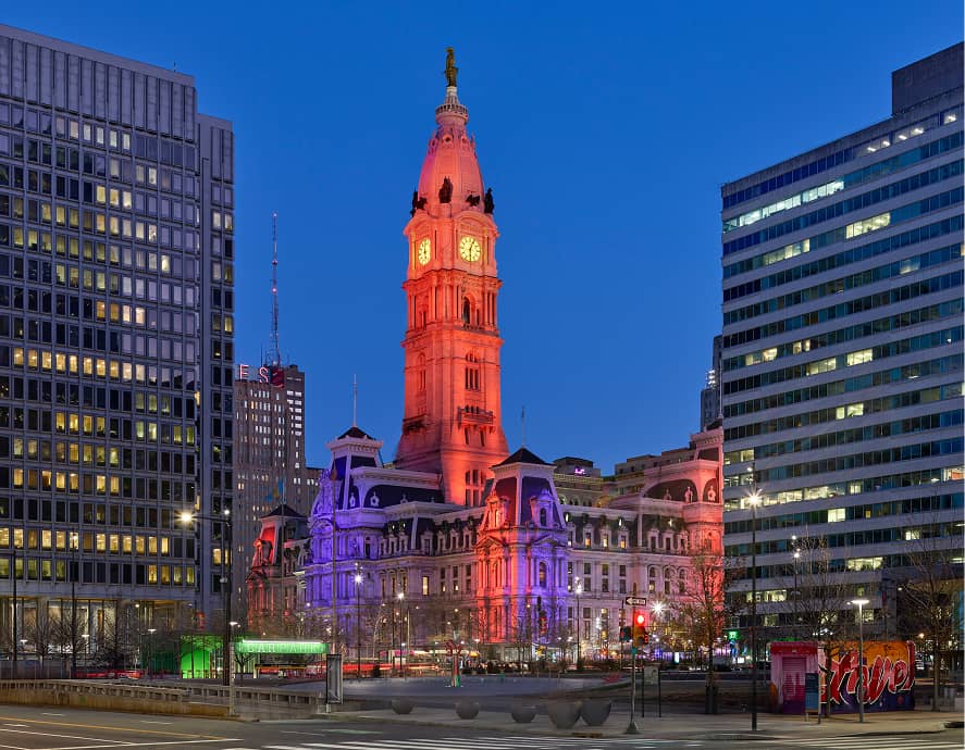 Philadelphia City Hall illuminated in red, white, and blue lights at dusk. The sky is clear, and modern buildings surround the historic landmark.