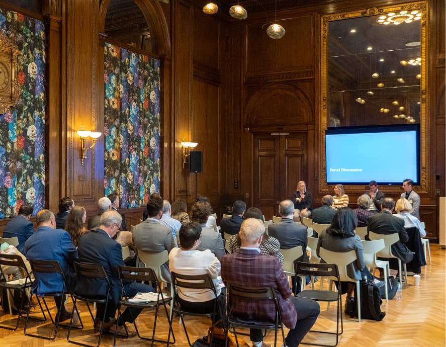 A group of people sit in a wood-paneled room with floral wall art, facing a panel discussion. The setting is formal with an air of focus.