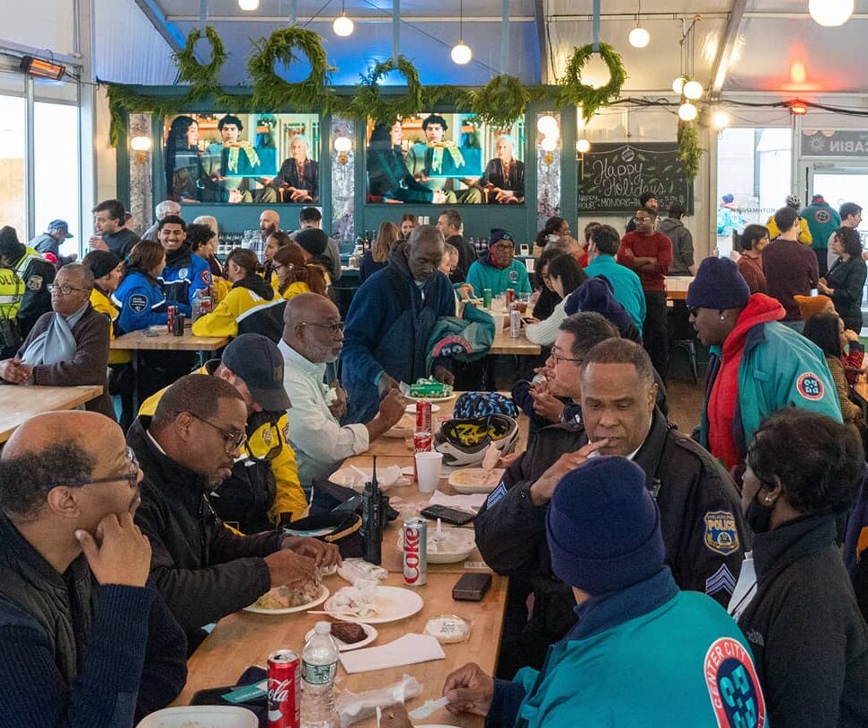 A diverse group of people enjoying a meal in a large, festive indoor space decorated with greenery. The atmosphere is warm and lively, with conversations and laughter.