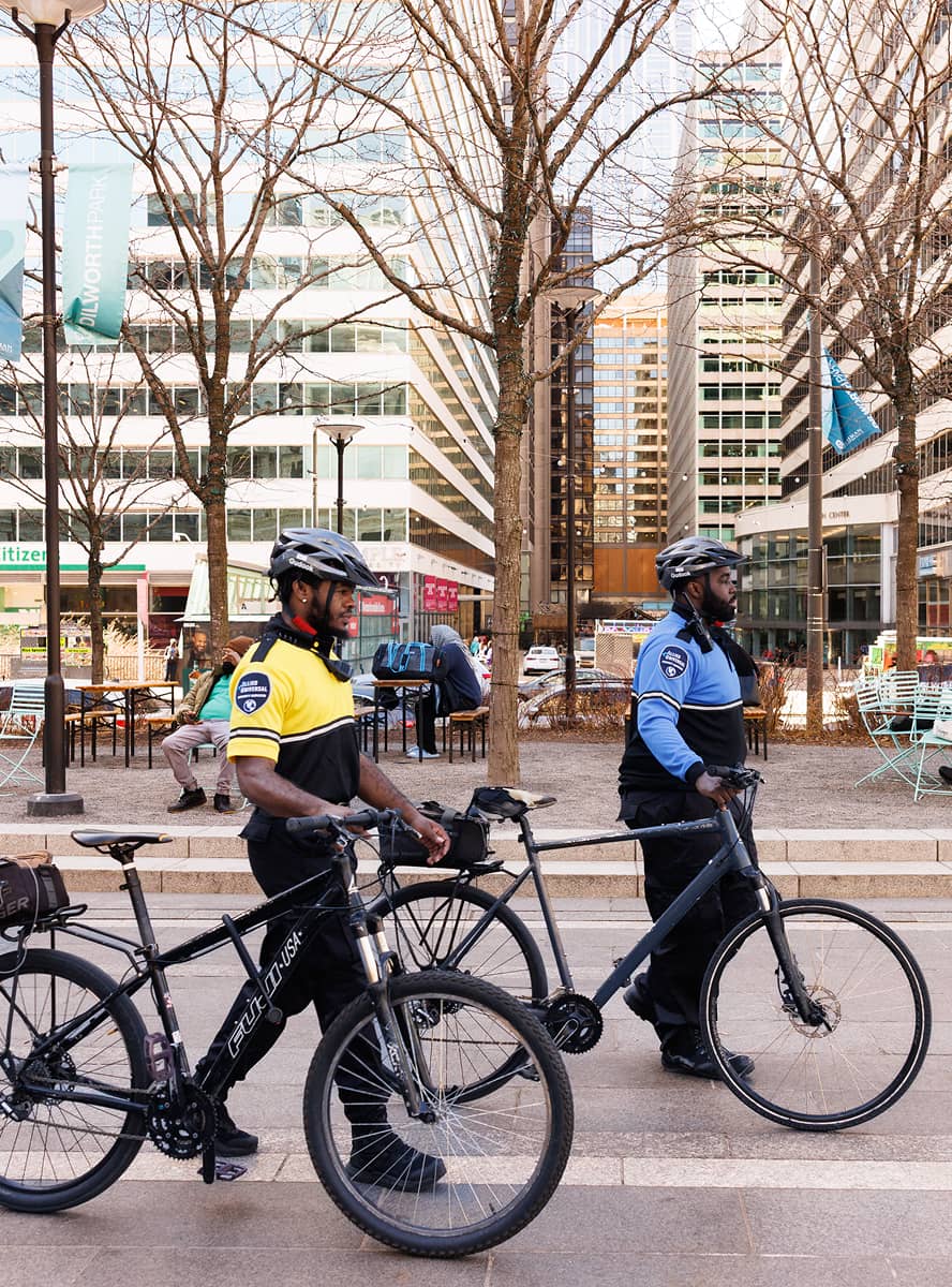 Two uniformed security personnel on bicycles patrol an urban plaza with modern buildings and leafless trees. The scene conveys duty and vigilance.