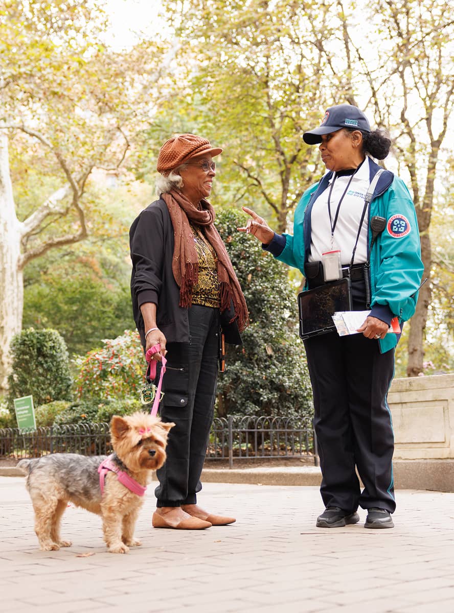 Two women converse in a park, one with a small dog on a pink leash. They appear friendly and engaged, surrounded by lush greenery on a sunny day.