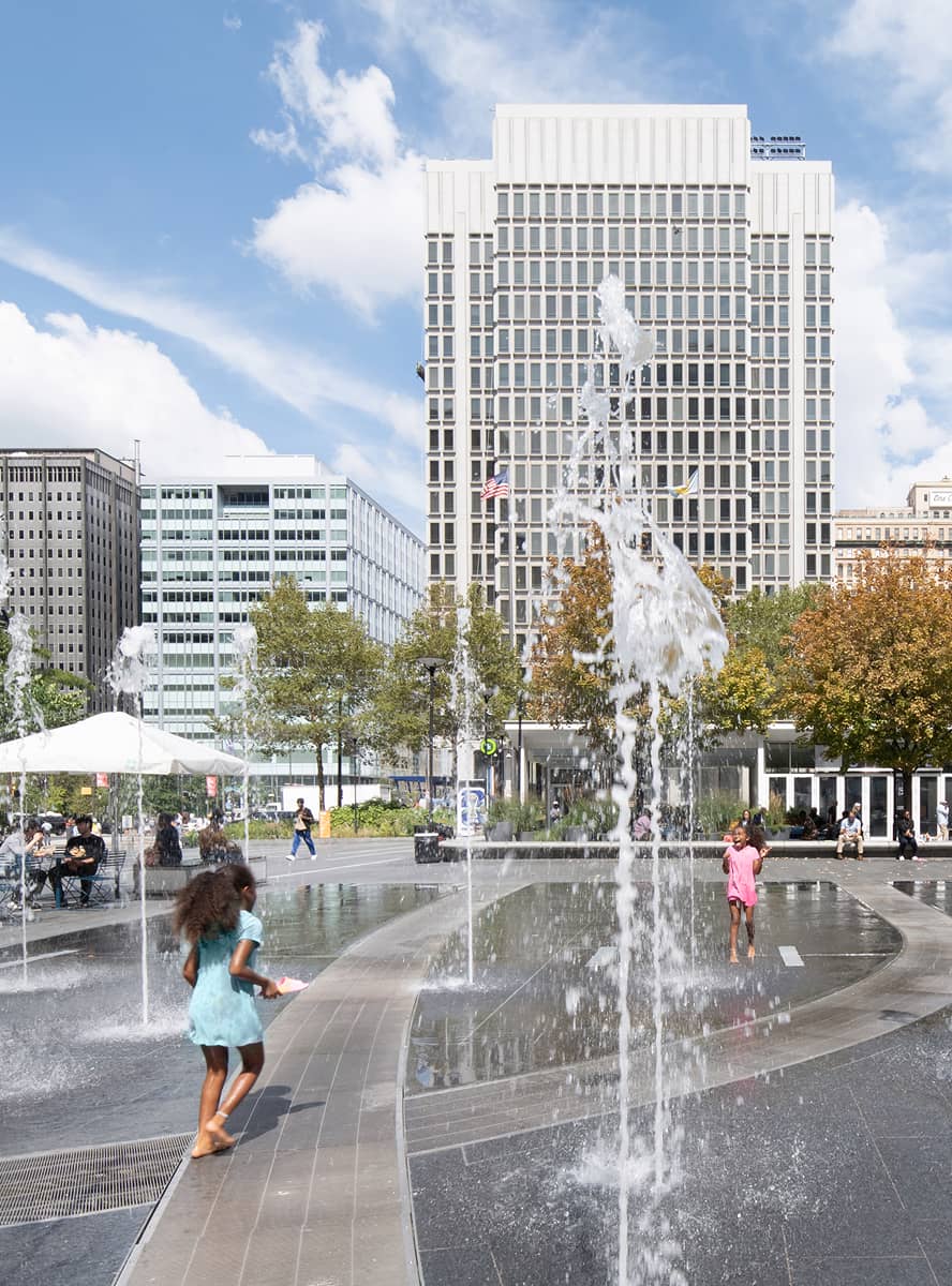 Children play joyfully in water fountains at a city plaza, surrounded by trees and modern buildings under a clear, blue sky.