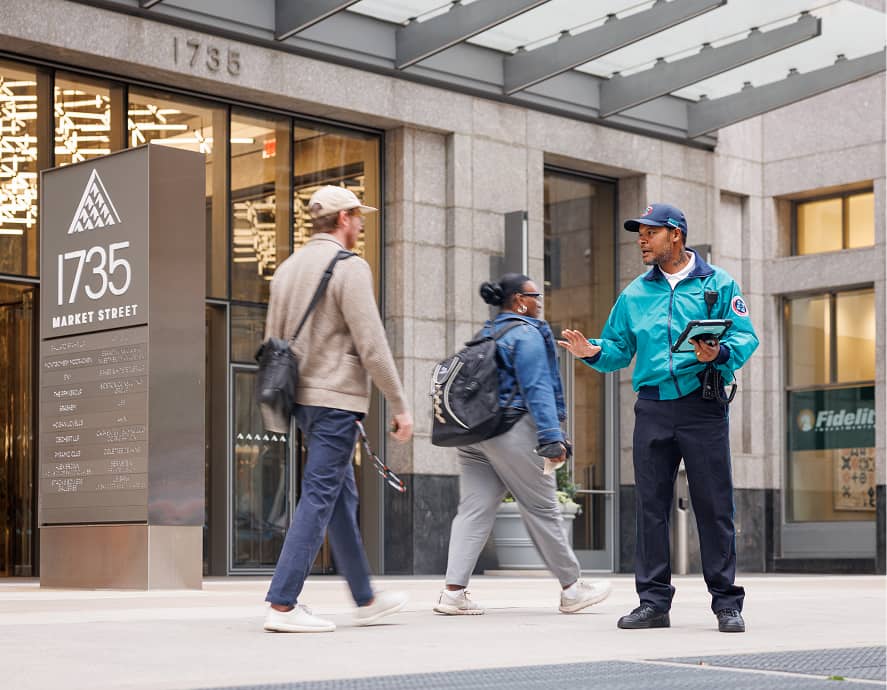 A security guard in a teal jacket holding a tablet gestures while speaking to pedestrians. Urban setting, overcast day, businesslike atmosphere.