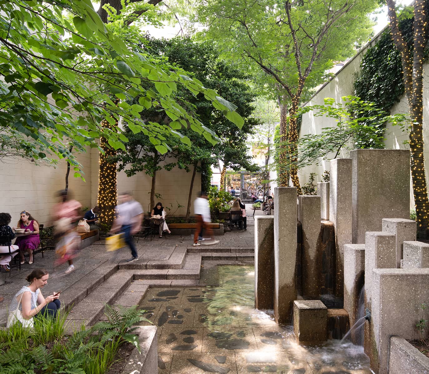Urban garden with lush trees and a stone fountain. People sit and walk, enjoying the peaceful atmosphere. Fairy lights wrap tree trunks, adding a warm glow.