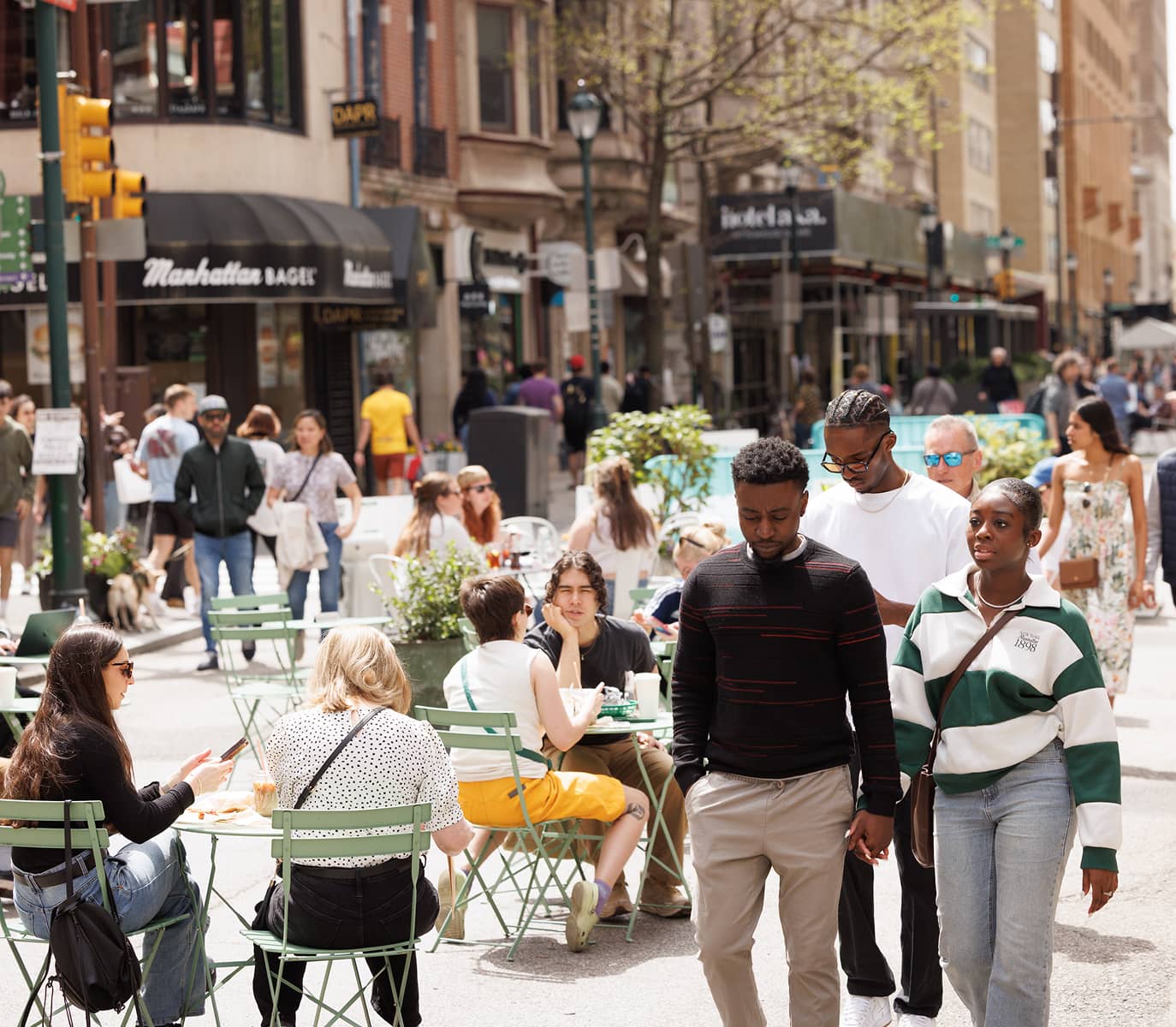 A lively city street scene with people dining at outdoor cafes. Three people walk by, surrounded by buildings, greenery, and a bustling, sunny atmosphere.