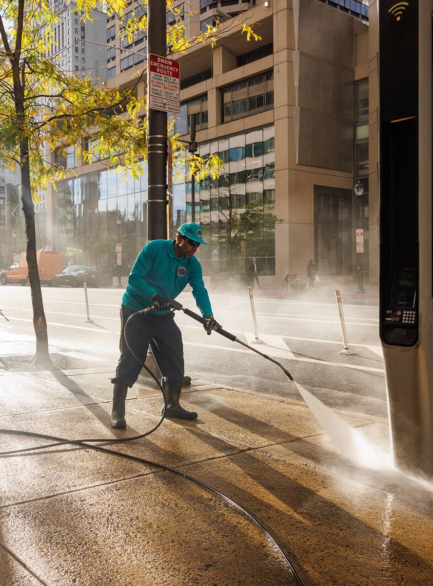 A worker in blue uniform pressure washes a sidewalk under sunlight in a city. Skyscrapers and a tree with yellowing leaves are in the background.