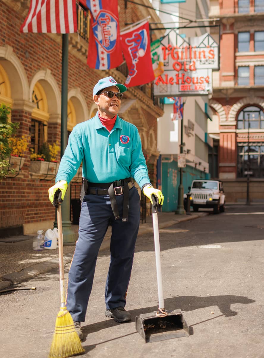 A person in a teal uniform is smiling and sweeping a street. They hold a broom and dustpan, with American flags and red-brick buildings in the background. The scene is bright and cheerful.