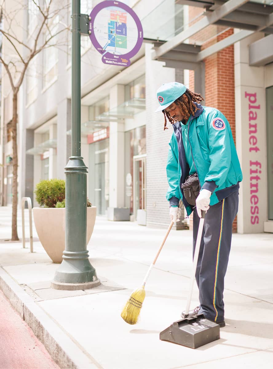 A person in a turquoise uniform and hat sweeps a city sidewalk with a broom and dustpan. The street is clean and empty, suggesting a quiet, orderly setting.